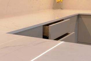 Modern kitchen corner with smooth white countertops and partially open drawers. Bright, clean, and minimalistic design with a sleek vase in the background.