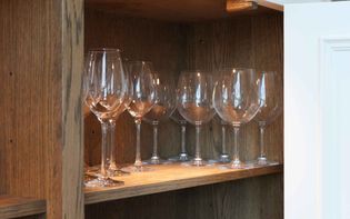 Eight clear wine glasses are neatly arranged on a wooden shelf in a cabinet. The light wood and clean glass create a tidy, elegant ambiance.