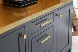 A grey shaker cabinet with gold handles and knobs is topped with a sleek brass counter. A black appliance is partially visible on the surface, adding elegance.