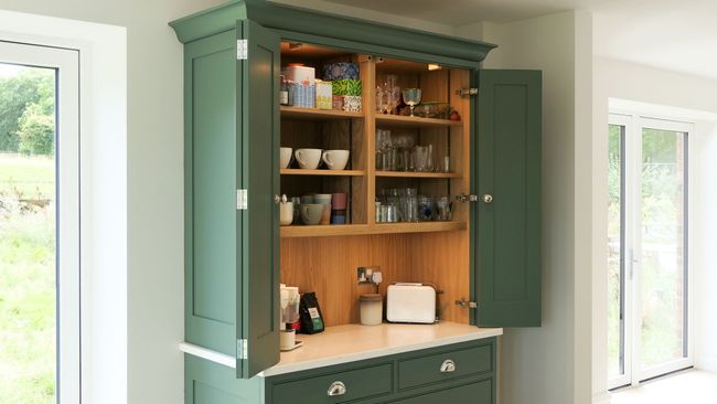 Green shaker kitchen dresser with open bi-fold doors, revealing shelves of mugs, glasses, and jars. A countertop holds a toaster and coffee supplies.