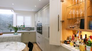 A Shaker kitchen featuring white cabinetry, a quartz wprltop, and a built-in bar with an assortment of liquor bottles and glassware.