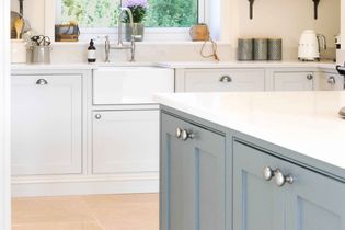 Bright kitchen with white and  cabinets, a farmhouse sink, and a light blue island. Sunlight filters through a window, illuminating a tidy, modern space.