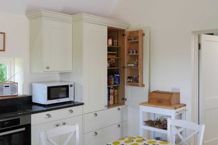 Bright shaker kitchen with an open pantry cabinet filled with food items and spices. Nearby, a microwave and breadbox sit on black countertops. Cozy atmosphere.