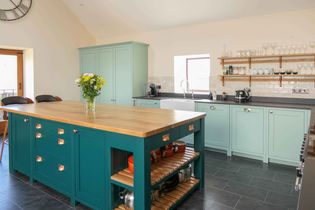 Bright kitchen with teal cabinets and large wooden island. Shelving holds glasses and mugs. A vase of flowers adds a touch of freshness.