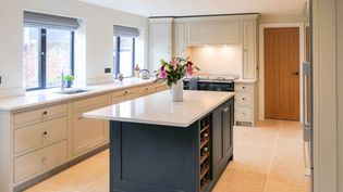 A shaker kitchen with cream cabinets and a dark island. Sunlight streams through two windows above the sink. A vase with flowers sits on the island.