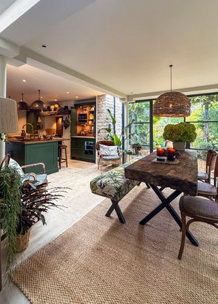 Bright modern dining and living area with a wooden table, pendant light, indoor plants, and large windows overlooking greenery.