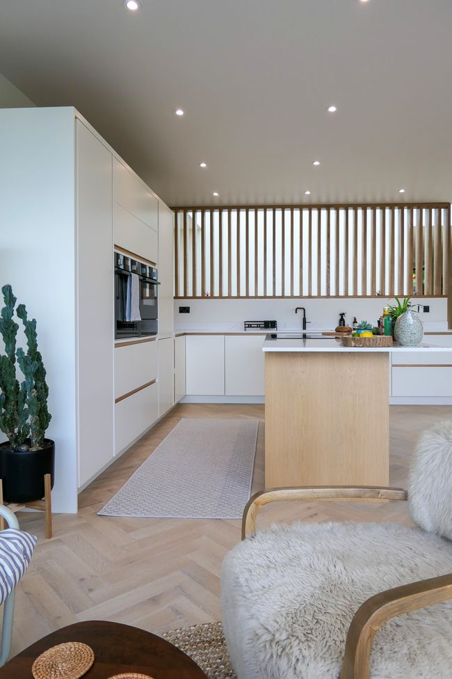 A modern kitchen featuring off white cabinets, integrated appliances, wooden accents, and a beige rug on a herringbone wood floor.