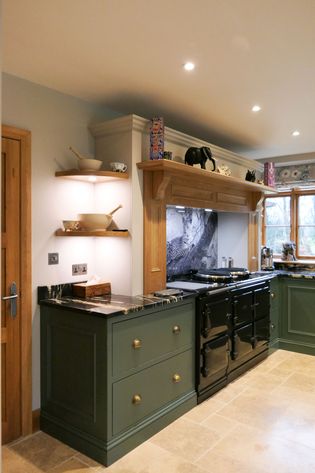 Warm kitchen with green cabinets and marble countertops. A black stove sits below a wooden mantel, with decorative bowls on shelves. Cozy and inviting.