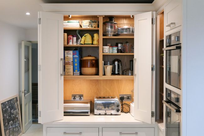 Open kitchen countertop cupboard reveals organized shelves with kitchen appliances, jars, and boxes. Lower shelves have a toaster and bread bin. Bright and tidy scene.