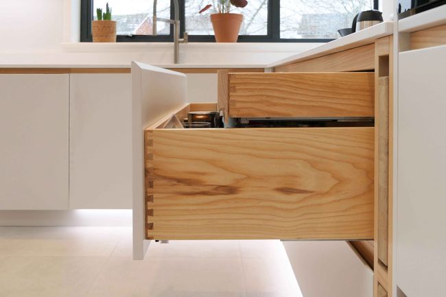 Modern kitchen with wooden drawers slightly open, revealing utensils. Sunlight streams through a window with potted plants on the sill. Sleek and minimalist design.