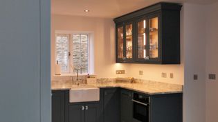 Shaker kitchen with dark cabinets, granite worktop, and a farmhouse sink. Glass cabinet displays crystalware. Bright, inviting space with under-cabinet lighting.