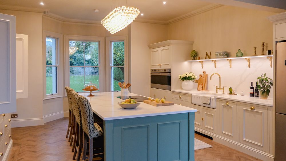 Large neutral and green Shaker kitchen with a central island, fruit, patterned bar stools, tall garden-facing bay windows, and a crystal chandelier.