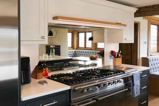 A shaker kitchen features a sleek stainless steel stove and oven, white cabinets, and a wooden beam. A cozy nook with windows and a bench is visible in the reflection from the mirrored splashback.