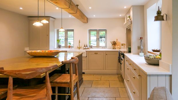 Warm, rustic Shaker kitchen with cream cabinets, a dark central island, brass fixtures, exposed beams, and large windows with a sink in-between them.