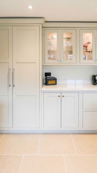 A shaker kitchen with light cabinetry, glass-paneled cupboards displaying glasses, a marble countertop, toaster, and coffee maker; warm, minimalist tone.