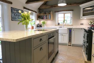 Rustic shaker kitchen with central island, exposed wood beams, and stone floor. A vase of flowers and books add warmth. Bright, cozy atmosphere.