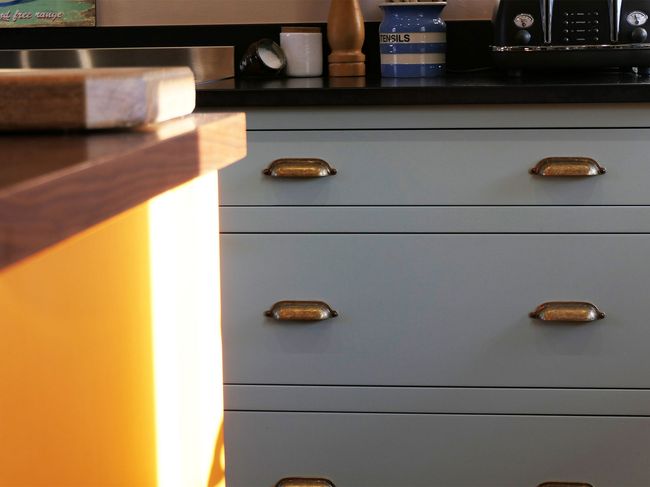 Sunlit kitchen scene with blue drawers, brass handles, and an orange counter. A pepper mill and utensil jar sit on a black countertop, creating a cozy atmosphere.