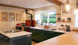 Country-style Shaker kitchen with dark cabinets, exposed wooden beams, a wood-burning stove, central garden-view window, and hanging glass lights.