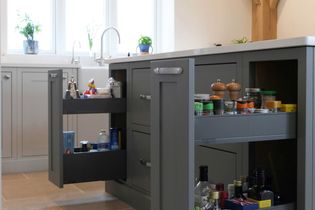 Shaker kitchen with open drawers on a dark grey island, displaying spices, condiments, and bottles. Sunlit, minimalist decor with potted plants by the window.