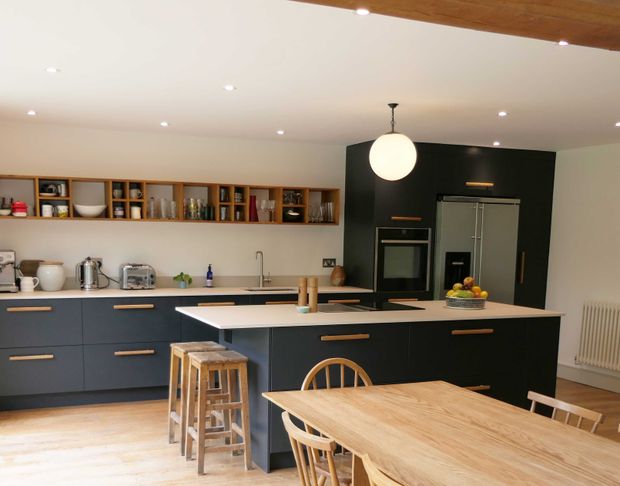 Modern kitchen with dark cabinets, open wooden shelving, a central island, fruit bowl, stools, and a wooden dining table.
