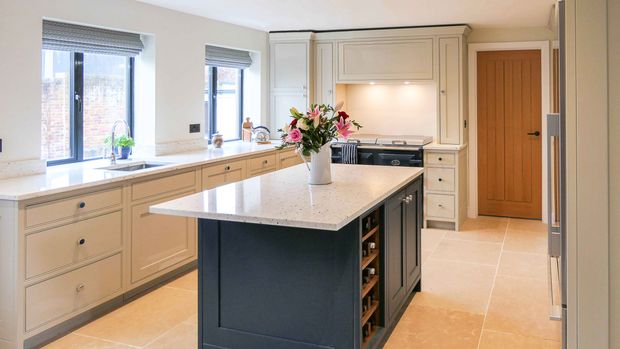 Shaker kitchen with cream cabinets, a dark blue island topped by a vase of colourful flowers, and windows overlooking a brick wall.