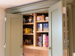 Open kitchen cabinet with light green doors revealing neatly organized shelves of dry goods, canned items, and mugs. Warm, tidy atmosphere.