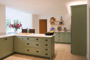 A shaker kitchen with green cabinets and light countertops. Bright flowers on the left, a basket of fruit on the island, and wooden shelves with decor on the right wall. Warm and inviting ambiance.