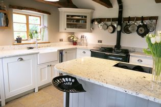 Rustic shaker kitchen with a white farmhouse sink, speckled worktops and hanging pots. A wooden beam ceiling adds warmth, while tulips on the island enhance coziness.