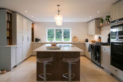 Large grey shaker kitchen with a central island, two perspex bar stools at one end, pendant lighting and a window above the sink over looking greenery in the garden.
