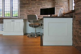 Rustic home office with stone walls, wooden floor, and a large window. Features a modern desk, curved monitor, and a gray swivel chair. Cozy and serene.