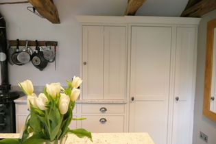 Cozy shaker kitchen with light grey cabinets, exposed wooden beams, and a hanging rack with pots. Fresh white tulips in a vase add a touch of elegance.