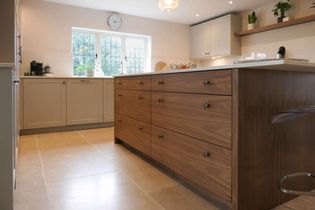 Modern kitchen with a central wooden island, beige cabinets, and light tiled flooring. A wall clock and plants on shelves create an airy, organized feel.