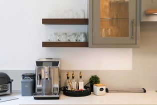 Shaker kitchen counter with espresso machine, tea jar, and condiments on a tray. Shelves hold clear glasses, and a small plant adds a touch of green.