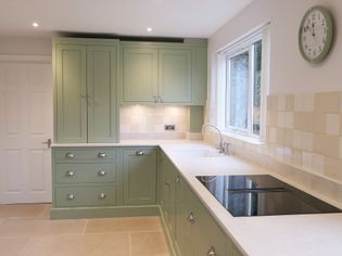 A shaker kitchen with green cabinets, sleek countertops, and beige tiled backsplash. A window offers natural light. A wall clock displays time.