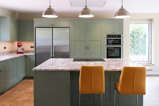 Shaker kitchen with green cabinets, stainless steel fridge, and built-in oven. Granite worktop island with mustard yellow chairs and pendant lights.