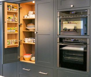Open kitchen pantry cabinets filled with spices and dishes beside two built-in ovens, one of which is open.
