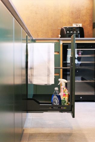 A close-up of a kitchen cabinet with a pull-out compartment holding cleaning supplies and a towel hanging on a rack.