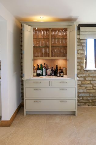 A off white shaker cabinet with open doors displaying glasses and various bottles on shelves, with a few drawers at the bottom, set against a stone wall.