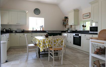 Traditional Shaker kitchen with white cabinets, various appliances on the black worktop and a dining table in the centre of the kitchen with a lemon patterned table cloth.