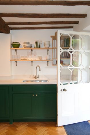 Charming kitchen with green cabinets, white countertops, and open shelves displaying dishes. A glass-paneled door and wooden ceiling beams add rustic elegance.