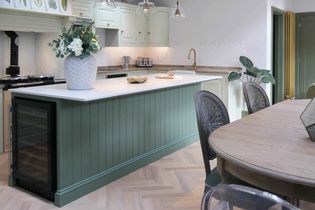 A shaker kitchen with a green island, white countertop, and vase of flowers. Light wood herringbone floor, dining table, and pendant lights create a cozy ambiance.