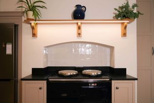 A cozy kitchen features a black oven with two rounded silver lids. Above, a wooden shelf holds a blue jug and green plants, creating a warm ambiance.