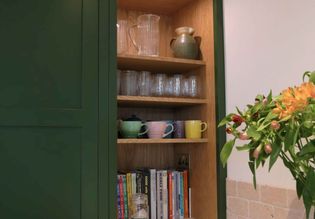 Open wooden kitchen cupboard with glass pitchers, mugs, and books. Bright flowers in the foreground add a cheerful touch to the scene.