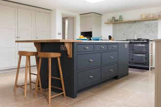 Shaker kitchen with a blue island featuring wooden stools, a hob in the background, and shelves with decorative pottery. Bright and inviting space.