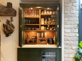 Countertop cupboard with various appliances, coffee, mugs and glasses, warmly lit with a drinks fridge and drawers beneath.