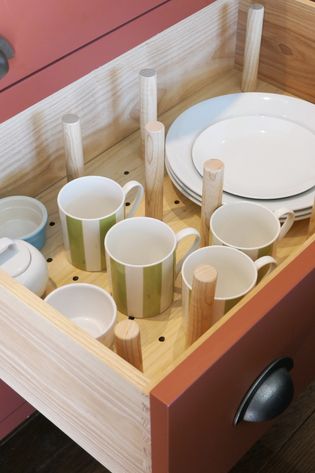 Wooden peg drawer holding white mugs, bowls, a lidded dish, and stacked plates separated by wooden dividers.