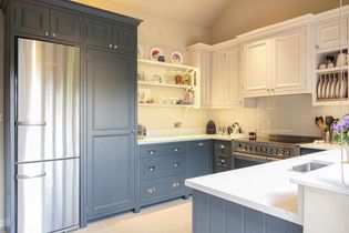 A shaker kitchen with navy blue lower cabinets and white upper cabinets. Stainless steel fridge, open shelves with dishes, and a built-in oven. Bright, tidy space.