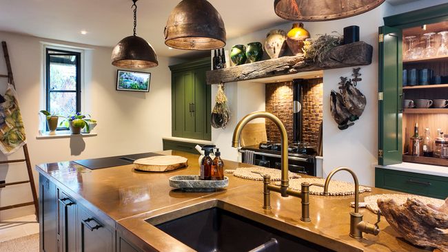 Warm rustic kitchen with a copper island, double sink, hanging pendant lights, open shelves, and green cabinetry.