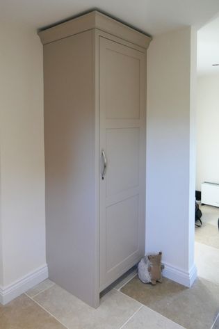 Tall beige cupboard with a brass handle, situated in a corner of a beige-tiled room. A small, plush hedgehog toy rests at the cupboard's base.