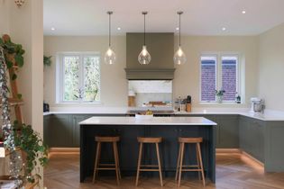 Shaker kitchen with a large island featuring three wooden bar stools. Pendant lights hang above, and green cabinets contrast with white countertops. Light streams through windows, adding warmth.